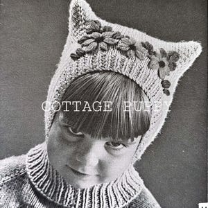以下が含まれることがあります： 花柄の装飾が施されたニットの猫の帽子をかぶった子供の白黒写真。帽子は尖った耳の形をしていて、子供の耳を覆っています。子供は真面目な表情でカメラを見つめています。