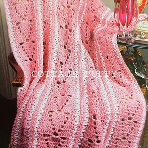 Puede incluir: Una manta de ganchillo rosa y blanca con un patrón de encaje. La manta está drapeada sobre una silla con un reposabrazos de madera. La manta tiene un borde festoneado.