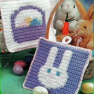 Könnte beinhalten: Zwei gehäkelte Topflappen mit Ostern-Motiv. Ein Topflappen zeigt einen lila-weißen Hasen, der andere einen lila-weißen Korb mit bunten Eiern.