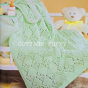 May include: A light green knitted baby blanket with a lacy pattern. The blanket is draped over a white crib with a stuffed teddy bear and a diaper bag.