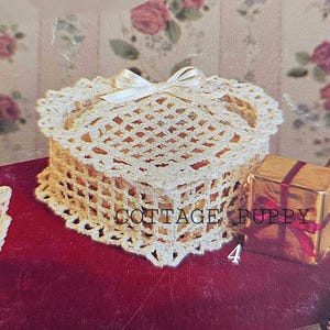 Könnte beinhalten: Eine gehäkelte herzförmige Schachtel mit einem weißen Satinband. Die Schachtel befindet sich auf einem roten Samtstoff und neben einer goldenen Geschenkbox mit einem roten Band. Der Text "COTTAGE PUPPY" ist auf dem Bild sichtbar.