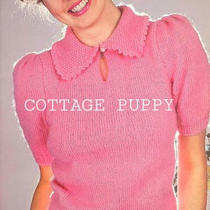 May include: A pink knitted sweater with a white collar and short sleeves. The sweater has a keyhole opening at the neckline and the text "COTTAGE PUPPY" is printed on the front.