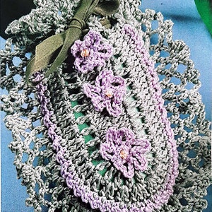May include: A crocheted lavender sachet with a green ribbon and three purple flowers. The sachet is on a blue background with two white heart-shaped objects.