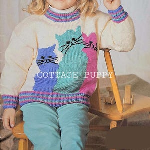 May include: A white knitted sweater with a colorful striped trim and three cat appliques. The cats are blue, green, and pink. The sweater is worn by a child sitting on a wooden chair. The text "COTTAGE PUPPY" is visible on the sweater.