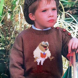 May include: A brown knitted sweater with a cartoon character design. The character is brown with a white shirt and orange tie. The sweater has brown buttons on the shoulders. The child wearing the sweater is wearing a brown and white patterned cap.