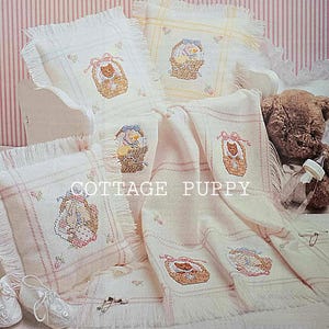 Puede incluir: Un conjunto de ropa de cama para bebés, que incluye almohadas y una manta, con diseños de punto de cruz de animales en cestas. Los artículos son de colores pastel suaves y tienen un ribete de flecos. El texto "COTTAGE PUPPY" es visible.