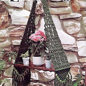 May include: A green macrame plant hanger with wooden shelf and beads hanging from the bottom. The hanger is hanging from a stone wall and has three potted plants on the shelf. The text "FRAME MAGIC" is visible at the top of the image.