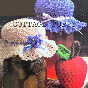 May include: Two crocheted jar covers, one in beige with a blue ribbon and a blue flower, and one in purple with a white ribbon. A crocheted red apple with a green leaf sits on a table with a crocheted doily.