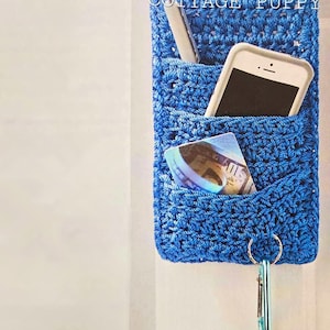 Könnte beinhalten: Ein blauer, gehäkelter Türknauf-Organizer mit Taschen für Schlüssel, ein Telefon und Karten. Der Organizer hängt an einer weißen Tür mit einem silbernen Türknauf. Der Text "COTTAGE PUPPY" ist auf dem Organizer sichtbar.  "Wenn Sie in Eile sind, um für Ihren geschäftigen Tag aus der Tür zu gehen, hilft Ihnen dieser Türknauf-Organizer, diese wichtigen Kleinigkeiten nicht zu vergessen."