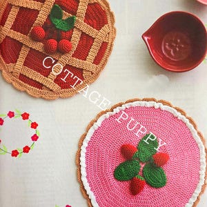 Op de afbeelding: Twee gehaakte placemats, een met een kersenpastei-ontwerp en de andere met een aardbeienontwerp. Beide placemats hebben een bruine gehaakte rand.