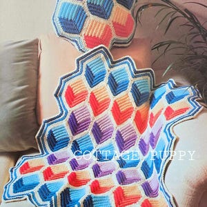 May include: A crocheted blanket and pillow with a three-dimensional design. The blanket and pillow are made with a variety of colors, including blue, purple, pink, and orange. The blanket is folded over a couch and the pillow is sitting on top of the blanket. The text "Three-Dimensional Design" is printed above the pillow.