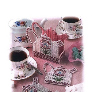 May include: A set of pink and white crocheted teapot-shaped coasters with floral designs. The coasters are arranged on a pink table with a teapot, teacups, and sugar cubes.