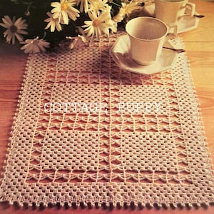 May include: Cream-coloured, rectangular crocheted table runner with an openwork design. The text "COTTAGE PUPPY" is visible on the runner. Two teacups and saucers are on the runner, with white daisies in the background.