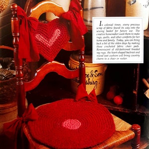 May include: A crocheted heart-shaped chair back cushion and a round crocheted chair seat cushion in red and pink. The cushions are on a wooden chair with a red fabric bow on each side.