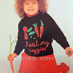 May include: A child wearing a black sweater with a colorful vegetable design and the text "I eat my veggies" is holding a carrot. The sweater is from Cottage Puppy.