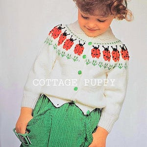 May include: A white knitted cardigan with a ladybug pattern on the front. The cardigan has a button closure and is worn by a young girl with curly brown hair. The girl is wearing a green corduroy skirt.