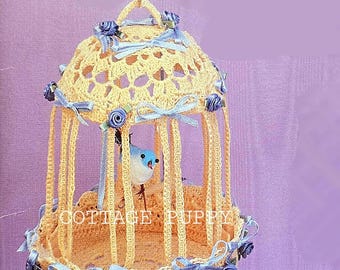 May include: A crocheted birdcage with a blue bird inside. The cage is decorated with blue and white ribbons and blue roses. The cage is hanging from a gold chain.