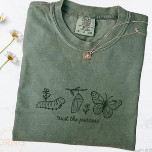 Peut inclure: T-shirt vert mousse avec un dessin d'une chenille, d'une chrysalide et d'un papillon, avec le texte "trust the process". Un collier en argent avec un petit pendentif est posé sur le t-shirt. Le t-shirt a une étiquette Comfort Colors.