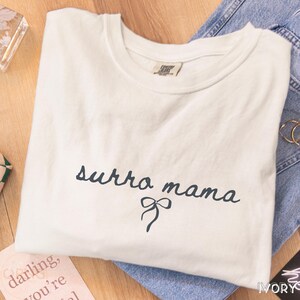 May include: Ivory-colored t-shirt with the text "surra mama" in a dark blue script font, accompanied by a bow graphic. The shirt is folded and placed on a wooden surface next to a pair of blue jeans.