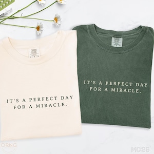 May include: Two Comfort Colors t-shirts are shown, one cream and one moss green. Each shirt features the phrase "IT'S A PERFECT DAY FOR A MIRACLE" in a simple font. The shirts are on a white surface with daisy accents.