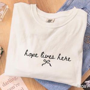 Peut inclure: T-shirt ivoire plié avec l'inscription manuscrite noire « hope lives here » et un motif de nœud. Le t-shirt est posé sur une surface en bois, à côté d'un jean bleu.