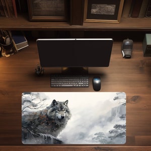 May include: A gray wolf with blue eyes is depicted in a watercolor style on a white desk mat. The wolf is standing on a rock with a waterfall and trees in the background. The desk mat is on a wooden desk with a computer monitor, keyboard, and mouse.
