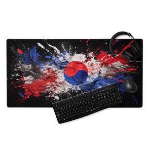 Pode incluir: Um tapete de rato preto com um design de pintura de respingos que apresenta a bandeira da Coreia do Sul. Também é mostrado um teclado preto e um fone de ouvido preto.