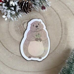Pode incluir: Um adesivo de um marmota marrom e branco segurando um ramo de folhas verdes. A marmota está em pé sobre uma superfície de madeira marrom claro.