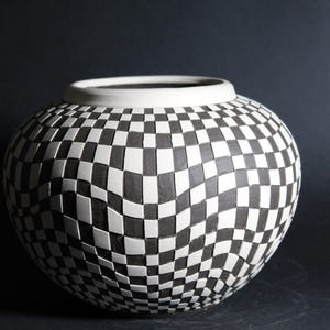 May include: A ceramic vase with a black and white checkered pattern. The vase has a round body and a white rim. The checkered design gives the vase a unique, textured appearance. The vase is a decorative item.