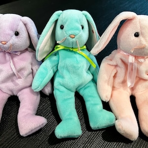 May include: Three plush bunny toys, one lavender, one teal, and one pink, all with long floppy ears and a bow tied around their necks.