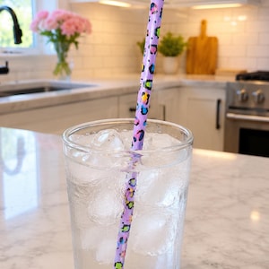 Op de afbeelding: Een helder glas gevuld met ijsblokjes en water, met een lange, rechte rietje met een lichtpaarse basis en een veelkleurig luipaardpatroon in tinten roze, blauw, groen en geel.
