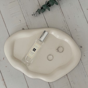 Może przedstawiać: Biała ceramiczna taca z falistym brzegiem, na której znajduje się butelka perfum Jo Malone London 'English Pear & Freesia Cologne' oraz dwa srebrne pierścienie. Taca leży na białym drewnianym blacie.