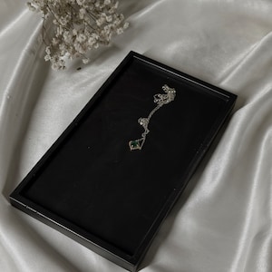 May include: A silver necklace with a green gemstone pendant on a black display tray. The tray is on a white satin fabric background.
