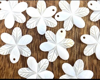 May include: A collection of white, flower-shaped pendants with a pearlescent sheen. Each pendant has five petals with etched details and a small hole for stringing. The pendants are arranged on a wooden surface.