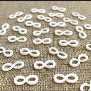 May include: A large quantity of white infinity symbol charms made from mother of pearl. The charms are scattered on a brown burlap surface.