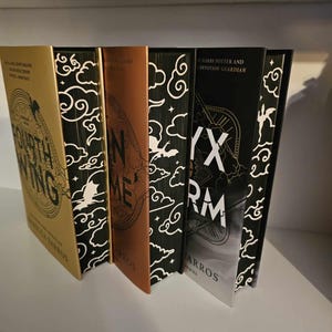 Puede incluir: Cuatro libros de tapa dura con cubiertas en blanco y negro que presentan un diseño de nubes. Los títulos son "Fourth Wing", "Iron Flame", "Six Crimson Cranes" y "Skyward", con el nombre del autor "Rebecca Yarros" en la parte inferior de cada libro.