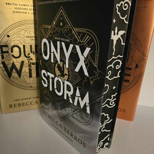 Könnte beinhalten: Ein schwarzes und goldenes Buch mit dem Titel "Onyx Storm" und den Worten "Brave the Dark" über dem Titel. Das Buch ist Teil einer Reihe namens "The Empyrean Series" von Rebecca Yarros. Der Buchumschlag zeigt ein stilisiertes Design eines Sturms mit Wolken und Sternen.