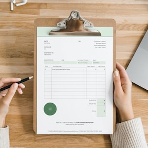 May include: A brown wooden clipboard holding a white invoice template with sage green accents. The document features fields for date, invoice number, customer ID, salesperson, job, payment terms, and a table for quantity, description, unit price, and line total.