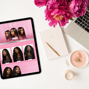 May include: A digital tablet displays a website with images of hair extensions. The tablet is surrounded by a laptop, notebook, pen, candle, and a small dish with paper clips. A bouquet of pink flowers sits above the tablet.