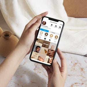 Op de afbeelding: Een persoon scrolt door een social media-app op hun telefoon. De app is Instagram en het profiel is voor een bedrijf genaamd "mystylecafe". Het profiel heeft een foto van een vrouw met bruin haar en een wit shirt. Het profiel heeft 1.000 volgers en 100 berichten. Het profiel heeft een raster met foto's in een neutrale kleurstelling.