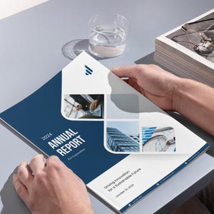 May include: A person holding open a corporate annual report. The cover is navy blue with the text "2024 ANNUAL REPORT" in white. The report features images of buildings and the text "Driving Innovation for a Sustainable Future". A glass of water and a stack of papers are also visible.