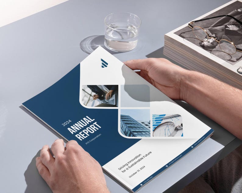 May include: A person holding open a corporate annual report. The cover is navy blue with the text "2024 ANNUAL REPORT" in white. The report features images of buildings and the text "Driving Innovation for a Sustainable Future". A glass of water and a stack of papers are also visible.