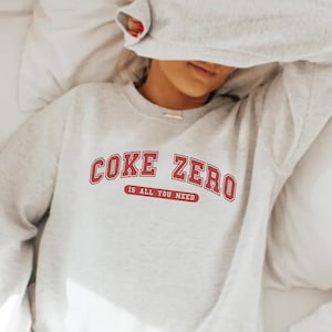 May include: A gray sweatshirt with the text "Coke Zero is all you need" in red lettering. The sweatshirt is on a white bed.