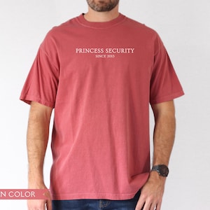 May include: Crimson-colored t-shirt with the text "PRINCESS SECURITY SINCE 2013" printed in white. The shirt has short sleeves and a crew neck. The model is wearing blue jeans and a black watch.