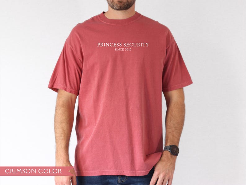 May include: Crimson-colored t-shirt with the text "PRINCESS SECURITY SINCE 2013" printed in white. The shirt has short sleeves and a crew neck. The model is wearing blue jeans and a black watch.
