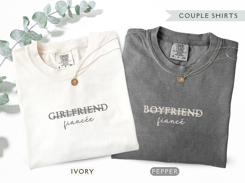 May include: Two t-shirts, ivory and pepper, with the words "Girlfriend fianc&eacute;" and "Boyfriend fianc&eacute;" printed on them. Each shirt has a gold necklace with a pendant. The shirts are labeled "Ivory" and "Pepper".