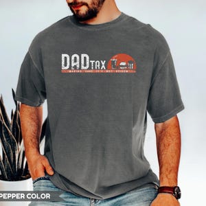 May include: Pepper color t-shirt with the text "DAD TAX" in white and red, along with a graphic of a burger, fries, and a drink. The text "MAKING SURE IT'S NOT POISON" is below the main text.