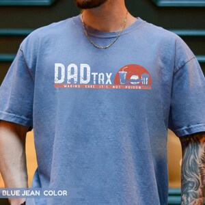 May include: Blue jean-colored t-shirt with the text "DAD TAX" in white block letters. Below the text is the phrase "MAKING SURE IT'S NOT POISON" in a smaller font. A graphic of a burger, drink, and fries is on the right.