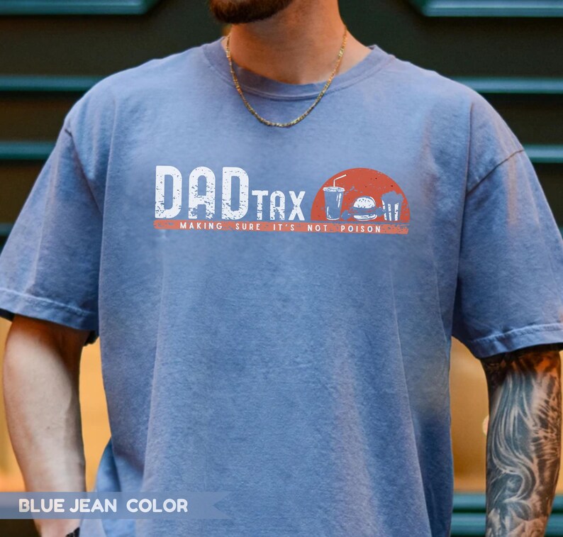 May include: Blue jean-colored t-shirt with the text "DAD TAX" in white block letters. Below the text is the phrase "MAKING SURE IT'S NOT POISON" in a smaller font. A graphic of a burger, drink, and fries is on the right.