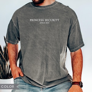 May include: Pepper color t-shirt with the text "PRINCESS SECURITY SINCE 2013" in white. The shirt is a faded gray color and has a vintage look. The model is wearing blue jeans and a brown watch.